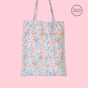 May include: A white tote bag with a floral pattern in shades of blue, pink, orange, and green. The bag has two long handles and a small logo that says "Tilly & Ashley Est. 2020".