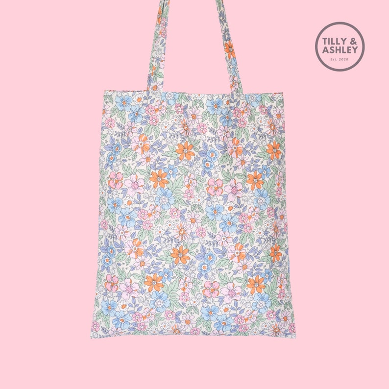 May include: A white tote bag with a floral pattern in shades of blue, pink, orange, and green. The bag has two long handles and a small logo that says "Tilly & Ashley Est. 2020".