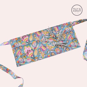 May include: A blue fabric apron with a floral pattern in pink, yellow, and white. The apron has two pockets and is tied with long blue fabric ties. The apron has four white plastic clothespins attached to it.