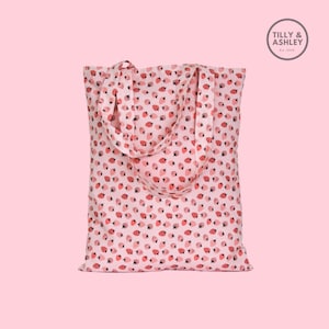 May include: A pink tote bag with a white background and a red strawberry pattern. The bag has two straps and the text "TILLY & ASHLEY Est. 2020" is printed on a white circle in the top right corner.
