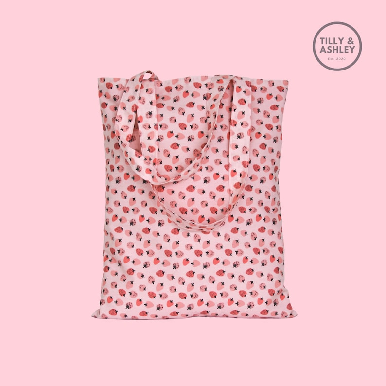 May include: A pink tote bag with a white background and a red strawberry pattern. The bag has two straps and the text "TILLY & ASHLEY Est. 2020" is printed on a white circle in the top right corner.
