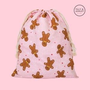 May include: Pink drawstring bag with a gingerbread man pattern. The bag has white drawstrings and a white label with the text "TILLY & ASHLEY Est. 2019".