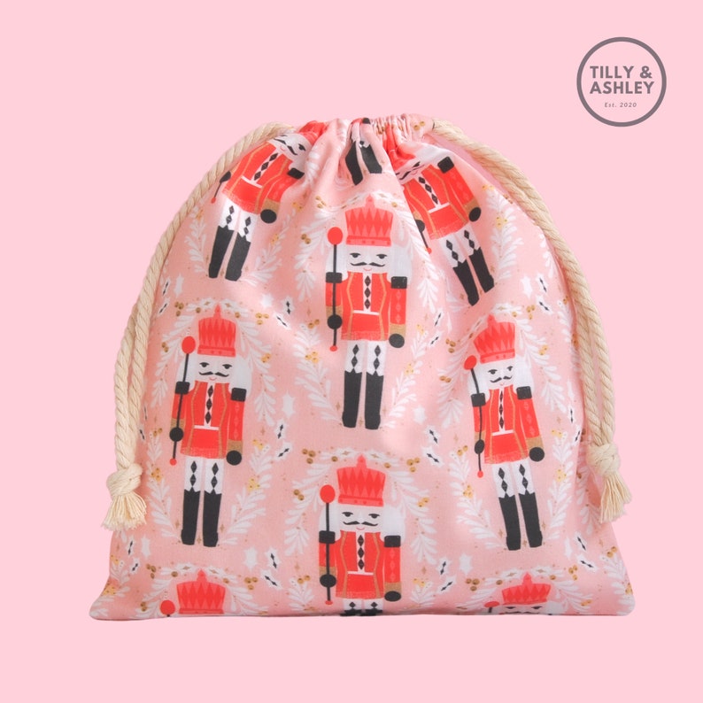 May include: A pink drawstring bag with a Nutcracker pattern. The bag features a repeating pattern of Nutcrackers in red and black uniforms holding a staff with a gold star on top. The bag has a white rope drawstring and the text "TILLY & ASHLEY Est. 2019" is printed on a gray circle in the top right corner.
