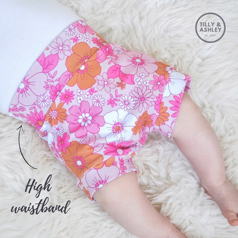 May include: Pink and orange floral print baby bloomers with a high waistband. The bloomers are made of soft, stretchy fabric and are perfect for a baby girl.