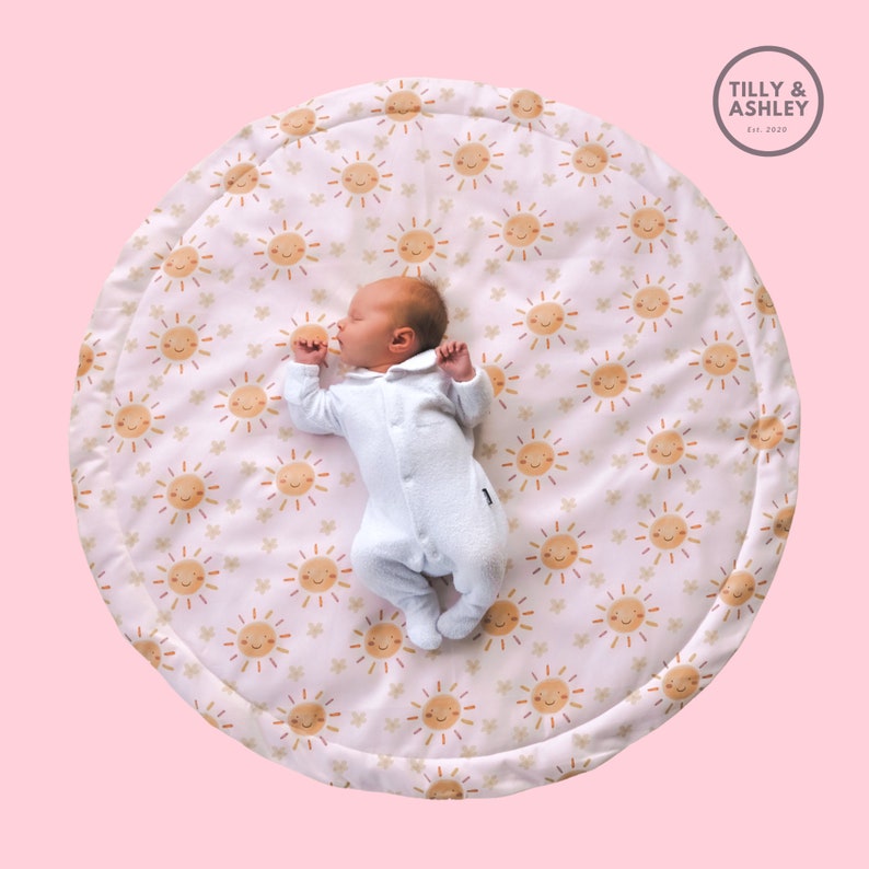 May include: A newborn baby in a white onesie lies on a round play mat. The mat features a repeating pattern of smiling suns and stars in shades of orange and tan on a cream background. The brand name "Tilly & Ashley" is visible.