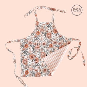 May include: A white apron with a floral print in shades of orange, pink, and brown. The apron has a gingham check pattern on the inside and ties at the neck and waist. The apron has the text "TILLY & ASHLEY Est. 2020" on the top right corner.