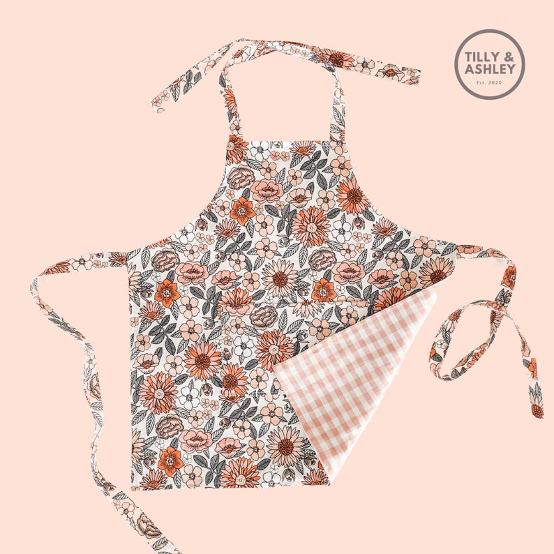 May include: A white apron with a floral print in shades of orange, pink, and brown. The apron has a gingham check pattern on the inside and ties at the neck and waist. The apron has the text "TILLY & ASHLEY Est. 2020" on the top right corner.