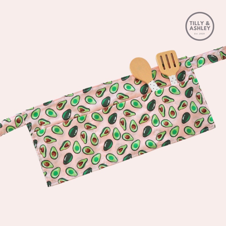 May include: A pink apron with a pattern of green avocados. The apron has two pockets and a long strap. Two wooden kitchen utensils are in the pockets.