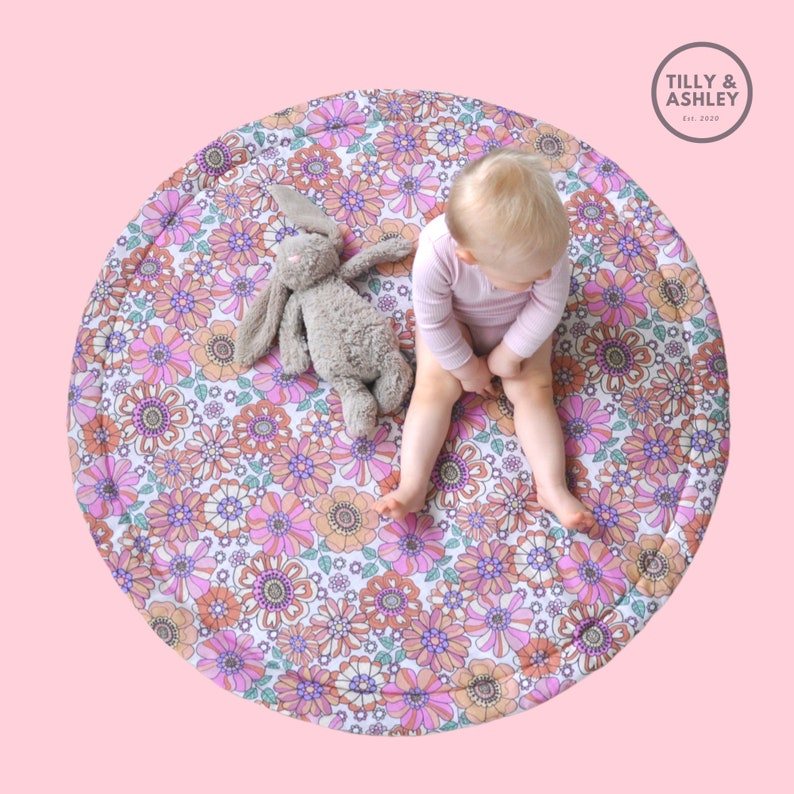 May include: A round, floral play mat with a vintage-inspired design in shades of pink, orange, and purple. A gray stuffed bunny and a baby in a pink onesie are on the mat. The mat has the text "Tilly & Ashley Est. 2020".