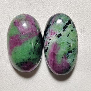 Könnte beinhalten: Zwei ovale, grüne, rosafarbene und schwarze Edelsteine. Die Edelsteine haben ein wirbelndes Farbmuster.
