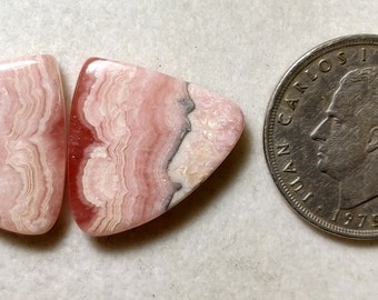 May include: Two pink and white triangular shaped cabochons of polished pink and white banded rhodochrosite. The cabochons are approximately the size of a US quarter.