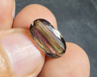 Puede incluir: Una gema de corte ovalado con inclusiones marrones, doradas y rojizas. La gema se sostiene entre los dedos, mostrando sus facetas pulidas y patrones internos. La superficie de la piedra refleja la luz, resaltando sus variaciones de color.