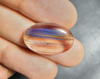 Puede incluir: Una piedra preciosa ovalada y translúcida con inclusiones horizontales iridiscentes que muestran un espectro de tonos azules, dorados y cobrizos. La piedra tiene una superficie lisa y pulida con un corte cabujón que resalta fibras internas.