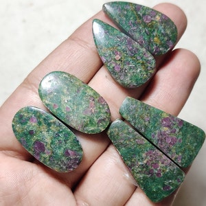 Könnte beinhalten: Eine Sammlung polierter Edelsteine in verschiedenen Formen, darunter Tropfen, Oval und Rechteck. Die Steine weisen eine grüne Grundfarbe mit lebendigen roten und rosa Einschlüssen auf, wodurch ein einzigartiges Muster entsteht. Die Edelsteine sind wahrscheinlich für die Schmuckherstellung bestimmt.