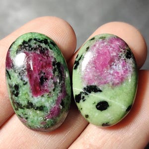 Könnte beinhalten: Zwei ovale, grüne, rosafarbene und schwarze Edelsteinkabochons. Die Steine haben eine marmorierte Optik mit grünen, rosafarbenen und schwarzen Wirbeln.