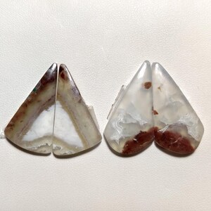 May include: Two pairs of polished agate stones. One pair features brown, white, and tan colours, while the other pair has a white base with red and brown inclusions. The stones are cut into a triangular shape.