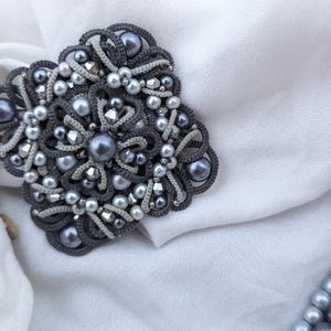 May include: A handcrafted, gray and silver brooch with a floral design. The brooch features a central large pearl surrounded by smaller pearls and silver accents. The intricate macrame-style design is set against a white fabric background.