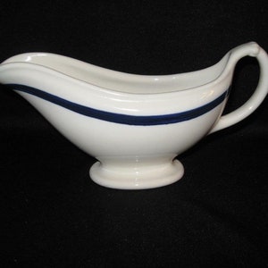 May include: White ceramic gravy boat with a blue band around the rim. The boat has a handle and a pedestal base.