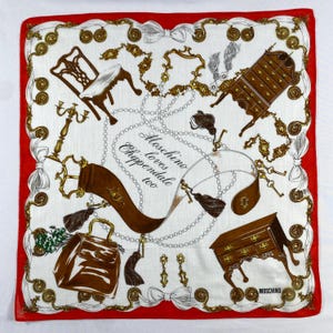 May include: A white Moschino scarf with a red border, featuring a repeating pattern of furniture, keys, and decorative elements in brown and gold. The text "Moschino loves Chippendale too" is printed on the scarf.