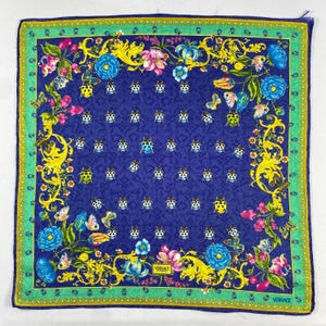 May include: A square silk scarf with a striking design. The scarf has a deep blue background decorated with ladybirds, butterflies, and floral patterns in pink, yellow, and blue. A green border frames the design, with the Versace logo visible.