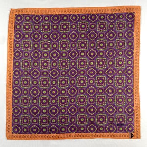 Könnte beinhalten: Ein quadratisches Textil mit einem sich wiederholenden geometrischen Muster in Lila, Grün und Gelb. Der orangefarbene Rand hat ein Maßbanddesign. Das Textil hat ein Etikett mit der Aufschrift "MOSCHINO".