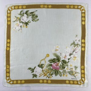 Può includere: Un fazzoletto quadrato azzurro con un bordo marrone ornato da medaglioni dorati. Il fazzoletto presenta composizioni floreali di fiori bianchi e rosa, con la scritta "Burberrys" stampata in basso a destra.