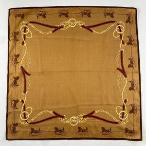 May include: A square, tan scarf with a brown border. The scarf features a repeating pattern of horse-drawn carriages and a gold chain and burgundy strap design. The word "CELINE" is printed on the border.