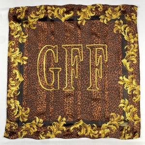 Könnte beinhalten: Quadratisches Seidentuch mit braunem Leopardenmuster und goldenen Akzenten. Der Schal hat die goldenen Buchstaben "GFF" und einen dekorativen goldenen Rand mit floralen Designs. Der Markenname "FRANCO FERRE" ist sichtbar.