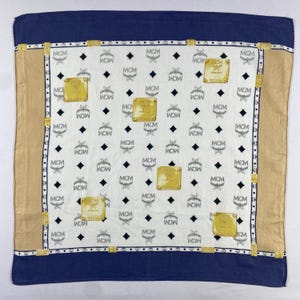 Peut inclure: Foulard carré avec un fond blanc orné d'un motif répété des lettres "MCM" et de losanges. Le foulard a une bordure bleu marine et des accents dorés. Les coins ont des patchs rectangulaires dorés.