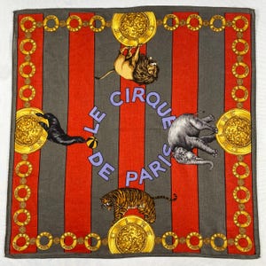 May include: A square scarf with a circus theme. The design features red and grey vertical stripes, gold medallions, and illustrations of circus animals, including a lion, elephant, tiger, and seal. The text "LE CIRQUE DE PARIS" is in the centre.