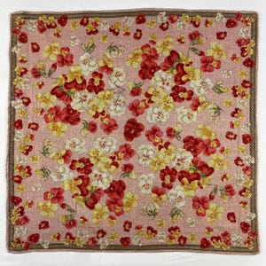 Può includere: Una sciarpa quadrata rosa con un motivo floreale. Il design presenta fiori rossi, gialli e bianchi sparsi sul tessuto. Un bordo marrone scuro con un motivo ripetuto incornicia i bordi della sciarpa.