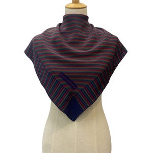 Pode incluir: Um lenço quadrado com um padrão listrado em tons de bordô, verde e azul, exibido em um manequim. O lenço tem uma borda azul escuro e o texto "pierre balmain".