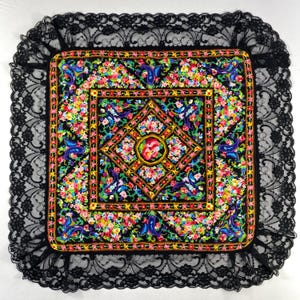 Peut inclure: Coussin carré décoratif à fond noir, orné d'un motif floral coloré et d'une bordure en dentelle noire. Le coussin présente un motif central en forme de losange avec des fleurs, encadré par une bordure florale carrée.