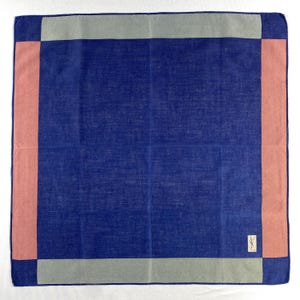 May include: A square, navy blue handkerchief with a light pink and gray border. The handkerchief has a small, rectangular tag in the lower right corner. The border is a textured, woven design.