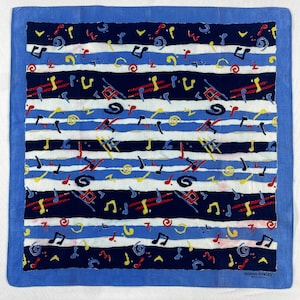 Peut inclure: Carré de tissu bleu et blanc à motifs, avec une bordure bleue. Le motif présente des rayures horizontales blanches, bleu marine et bleu clair, ornées de notes de musique et de formes abstraites rouges, jaunes et noires. Le texte "SONIA RYKIEL" est visible en bas à droite.