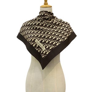 May include: A brown and cream silk scarf with a repeating pattern. The scarf has a dark brown border and the text "Christian Dior" is visible. The scarf is draped over a mannequin.