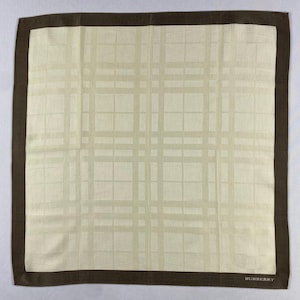 May include: A square, cream-coloured handkerchief with a brown border and a plaid pattern. The word "BURBERRY" is printed in the bottom right corner. The handkerchief is made of a soft fabric.