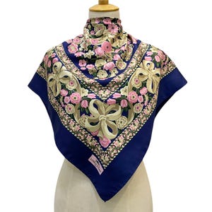May include: A square silk scarf with a floral pattern and a navy blue border. The scarf features pink and white flowers, beige bows, and a brand label. The scarf is displayed on a mannequin.