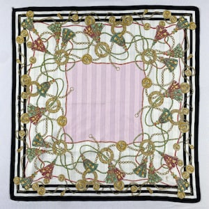 Könnte beinhalten: Quadratisches Tuch mit weißem Hintergrund und einem rosa gestreiften Mittelteil. Es zeigt goldfarbene Ketten, Münzen und Quasten in Grün und Rot. Ein schwarz-weiß gestreifter Rand umrahmt das Design. Dekoratives Accessoire.
