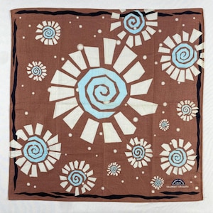 Könnte beinhalten: Quadratischer brauner Stoff mit einem sich wiederholenden Muster aus weißen und hellblauen Spiraldesigns. Der Stoff hat einen schwarzen Rand und kleine weiße Punkte. In der unteren rechten Ecke steht der Text "tsumori chisato".