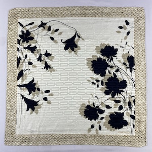 Peut inclure: Foulard carré en soie avec un motif floral. Le foulard présente un centre crème avec un motif géométrique et des accents floraux noirs. La bordure est beige texturé. La marque "NINA RICCI" est visible.