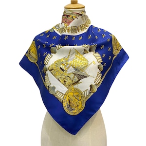 May include: A square silk scarf with a nautical theme. The scarf features a central design of a sailing ship in yellow and gold, framed by a white border and a deep blue background. The scarf is adorned with fleur-de-lis and shell motifs.