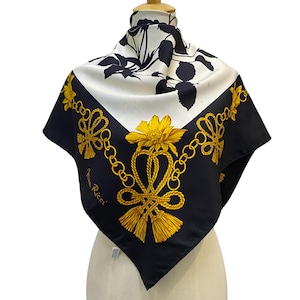 Pode incluir: Um lenço quadrado de seda com um padrão floral preto e branco e uma borda preta com desenhos florais e de correntes douradas. O lenço tem uma flor amarela no centro e a marca "Nina Ricci" é visível.