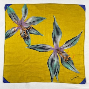 May include: A square, yellow scarf with a floral design. Two large flowers with purple centers, teal petals, and orange stamens are printed on the scarf. The corners have blue accents, and the designer's signature is in the bottom right corner.