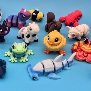 May include: A selection of small, brightly coloured animal figurines, such as a hedgehog, cat, ladybug, shark, frog, and axolotl, displayed on a blue background. The figurines are made of a smooth material and have detailed designs.