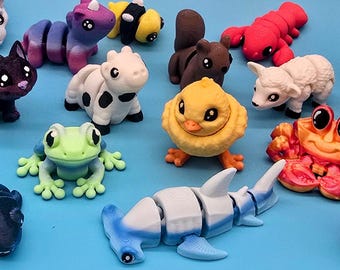 Op de afbeelding: Een verzameling kleurrijke, miniatuur dierenbeeldjes, waaronder een egel, kat, lieveheersbeestje, haai, kikker en axolotl, gerangschikt op een blauw oppervlak. De beeldjes zijn gemaakt van een glad materiaal en hebben gedetailleerde ontwerpen.