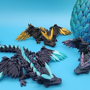 May include: Three origami dragons, one yellow, one black with blue accents, and one black with red and blue accents, are arranged on a blue surface. A blue and purple origami dragon egg is in the background.