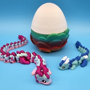 May include: Three colorful dragon toys, one in pink and blue, one in blue and green, and one in a curled up position. The dragons are all made of plastic and have a segmented body. The dragons are sitting on a blue surface in front of a white egg with a green and red base.