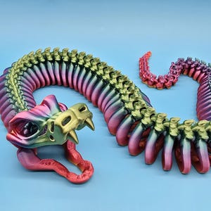 May include: A multi-coloured articulated dragon toy in shades of pink, green, and gold. The dragon is posed on a light blue surface, with its head and tail curved. The dragon's body is made of many connected segments.