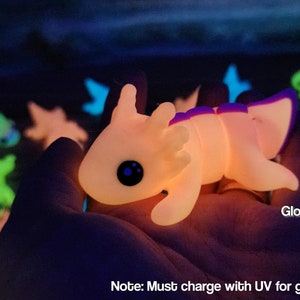 May include: A glow-in-the-dark axolotl figurine in pink and magenta. The figurine is made of a soft, flexible material and has a cute, cartoonish appearance. "Glow Pink / Magenta" and "Note: Must charge with UV for glow. See description." are written below the figurine.